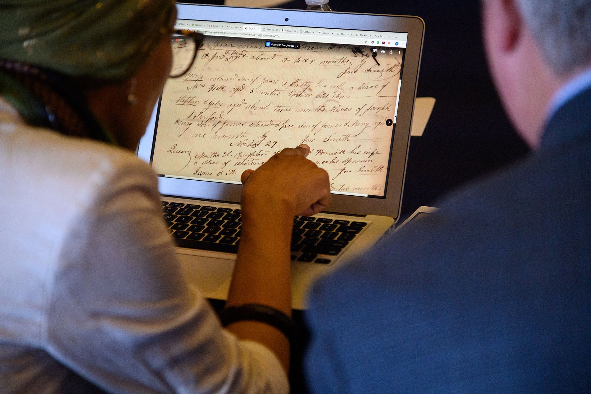 Two people read a handwritten historical document on a laptop computer screen.