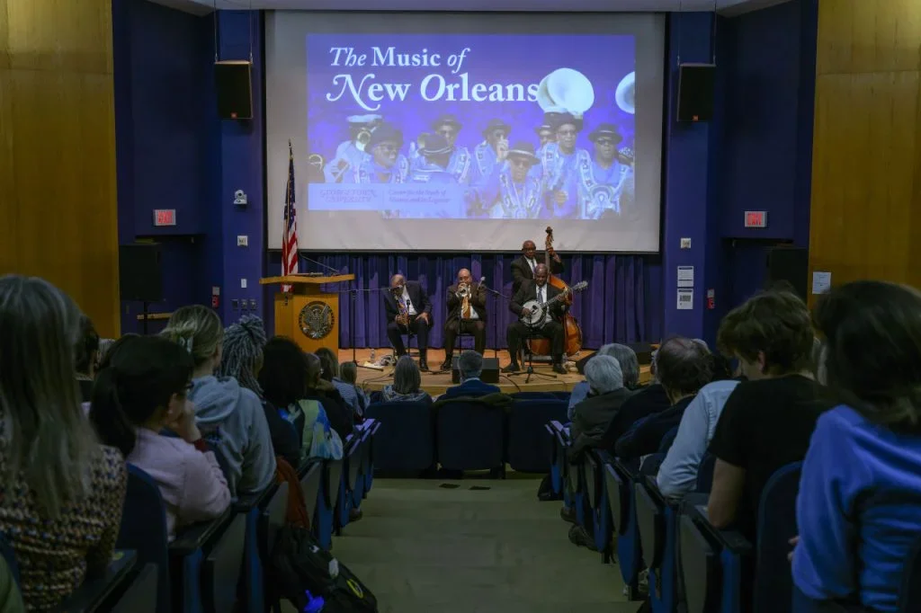 Four black musicians – clarinet, trumpet, banjo, and upright bass – perform on a stage in front of an audience. An image of other musicians, titled 