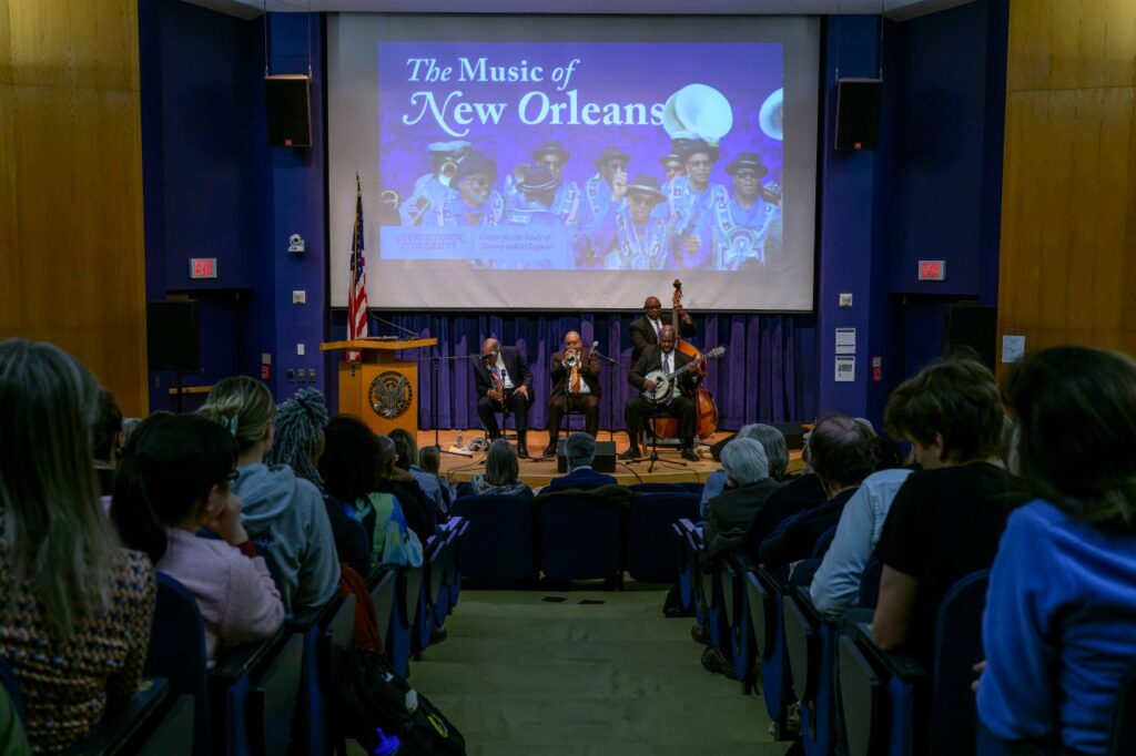 A four-piece band performs for a seated audience. Behind them is a projection screen that reads "The Music of New Orleans".