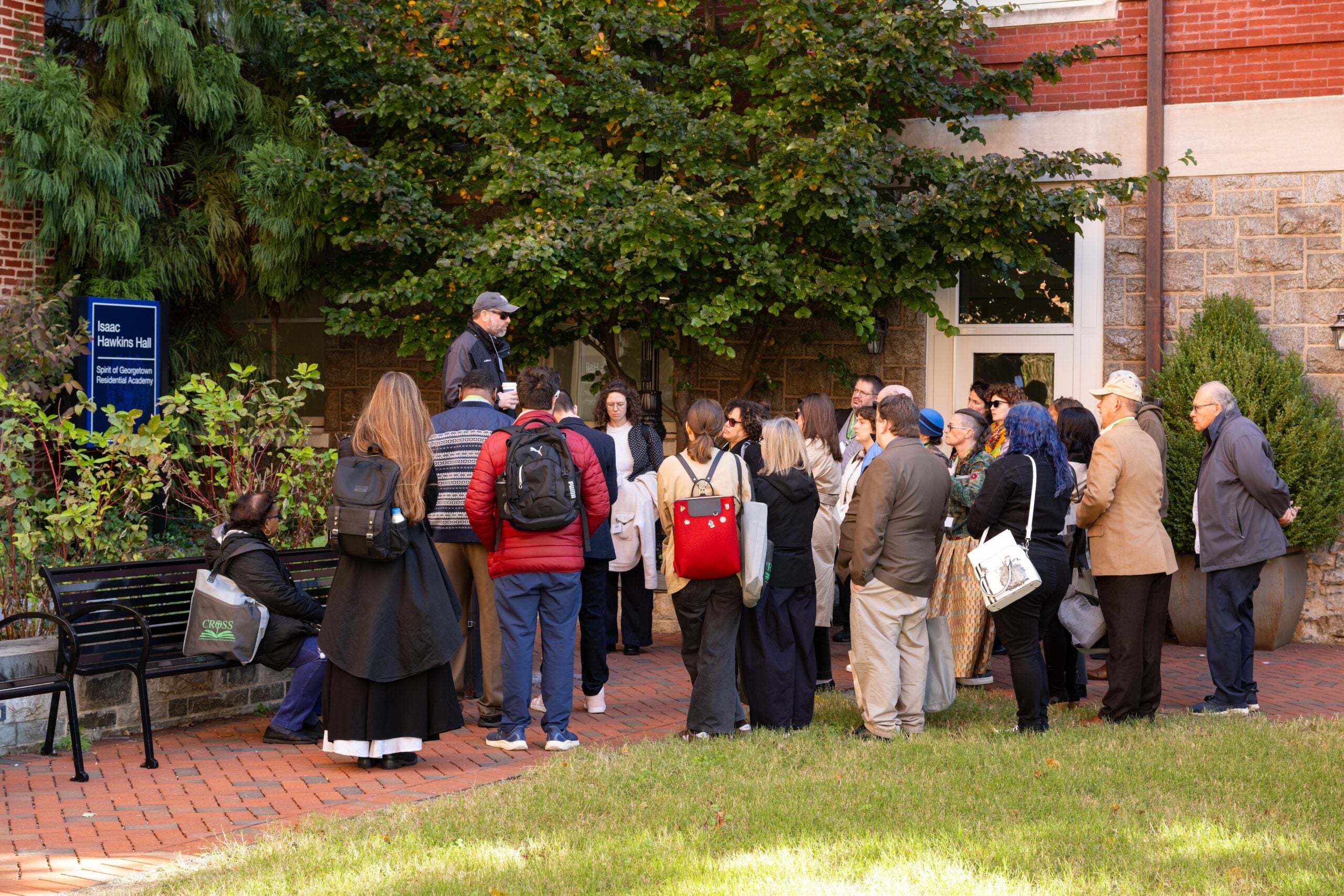 Adam Rothman, a white man wearing a jacket, sunglasses, and hat, stands on a bench and speaks to a group of walking tour participants. They are outside a brick and stone building, with a sign beside it that reads Isaac Hawkins Hall.