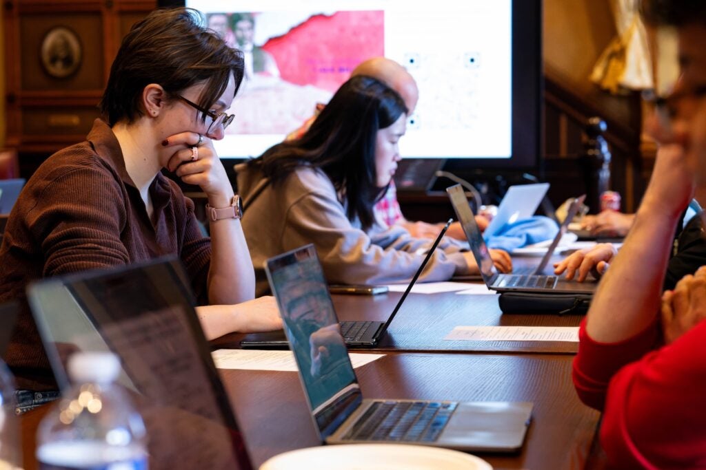 Two volunteers sit at a wooden table in a conference room and stare intently at laptop screens.