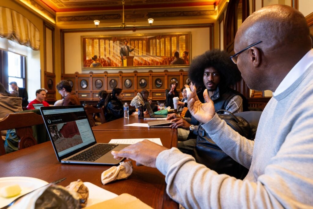 Two black men converse while sitting in a conference room, conducting research on their laptops.