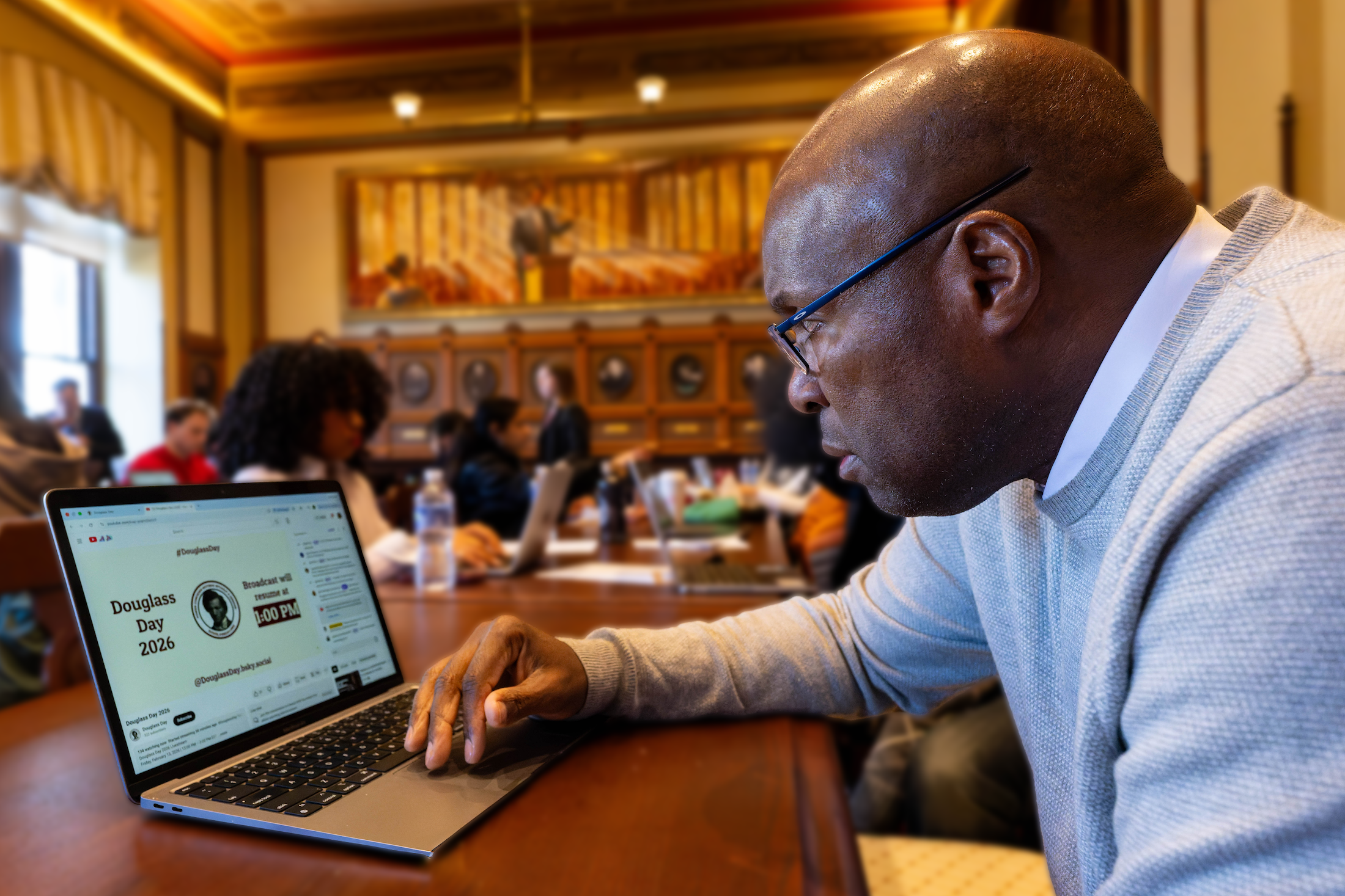 A black man wearing glasses sits at a wooden table in a conference room with other people. He is working on a laptop, whose screen reads Douglass Day 2026 with an illustration of Frederick Douglass.