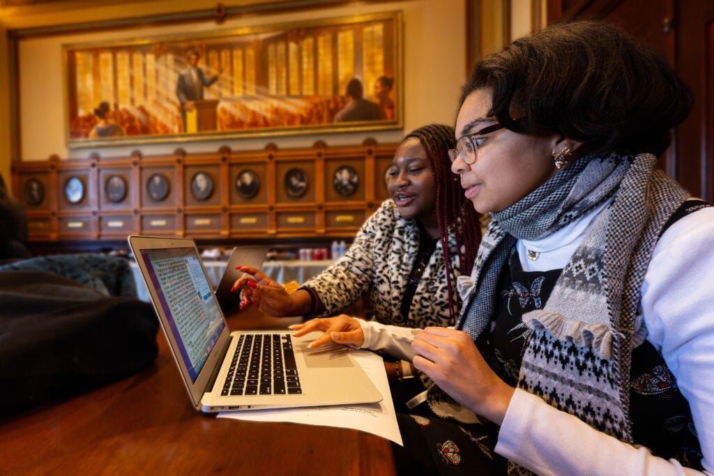 Two volunteers sit together at a wooden table in the Philodemic Room, collaborating as they look at the same laptop screen.