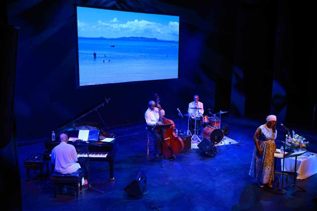 Michaela Harrison and her band, consisting of a pianist, a bassist, and a drummer, perform on a large stage. Behind them is a screen showing a beach scene.