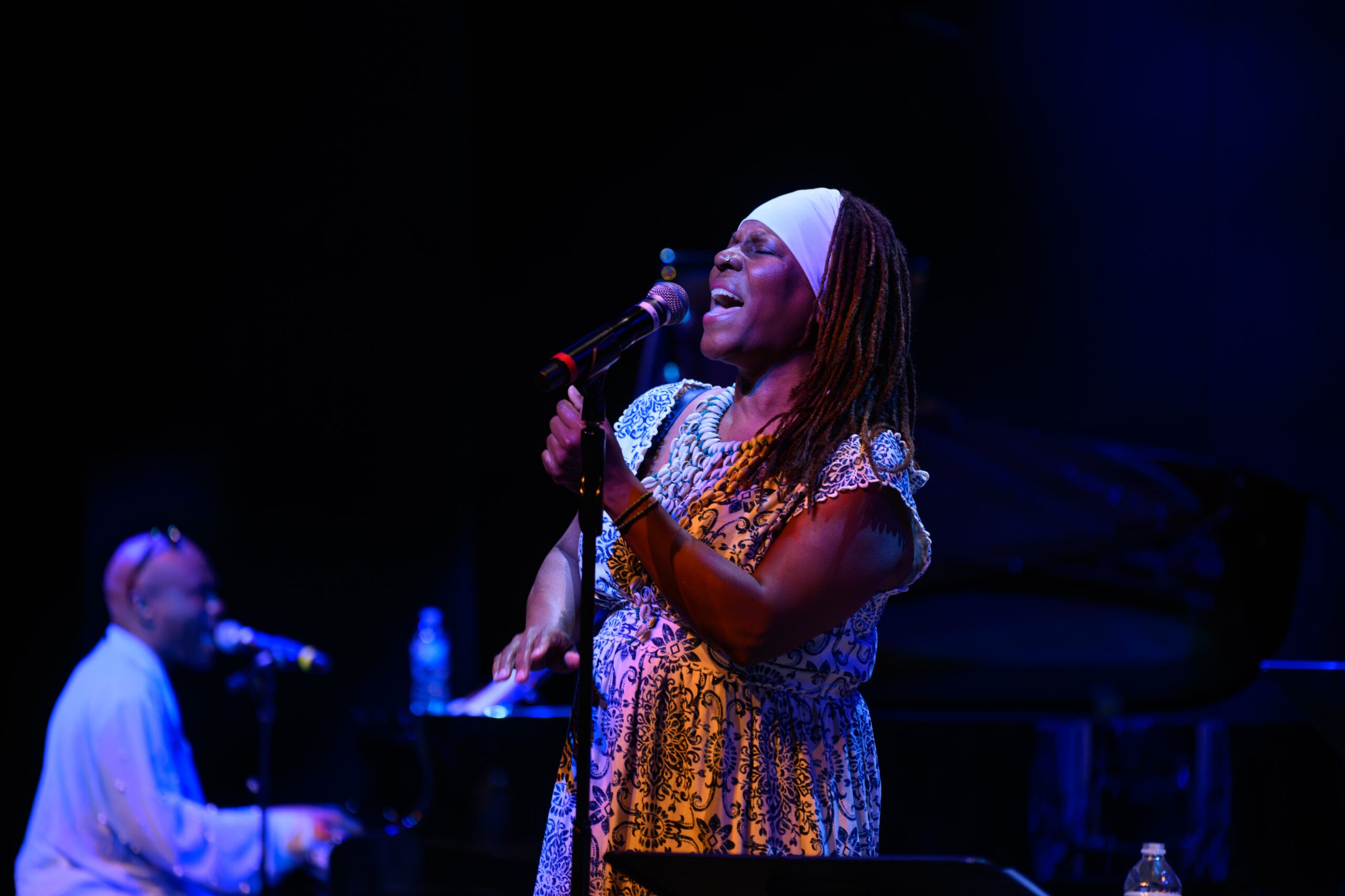 Michaela Harrison, a black woman with long braids, wearing a blue and white dress, sings at a microphone on stage. Behind her is a pianist, also singing.