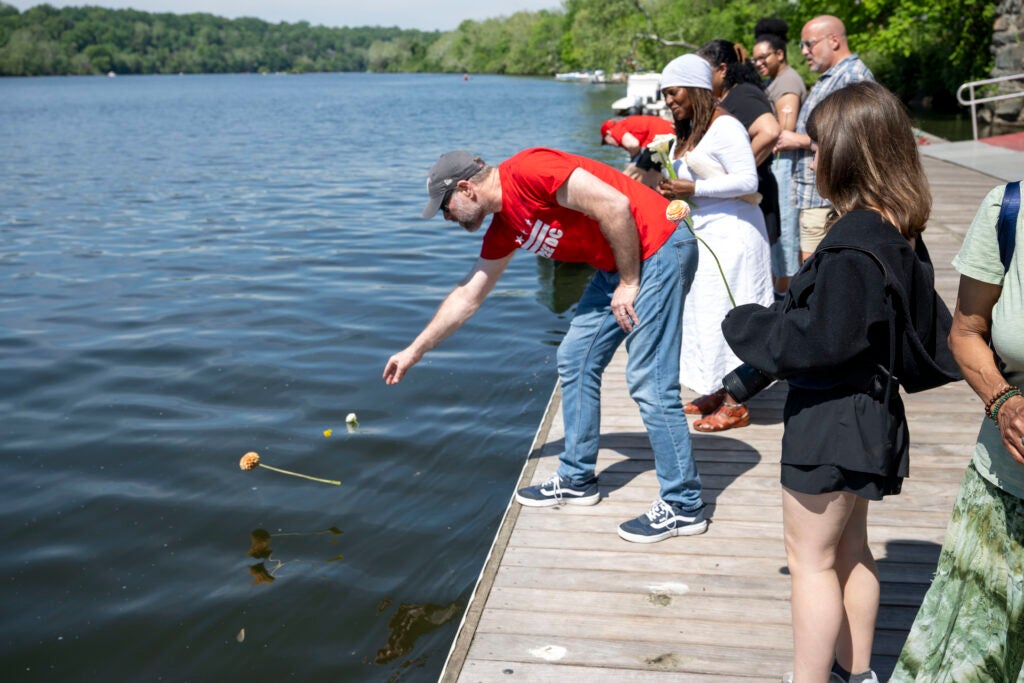 A group of people stand on a wooden pier. One man throws a small flower into the water.
