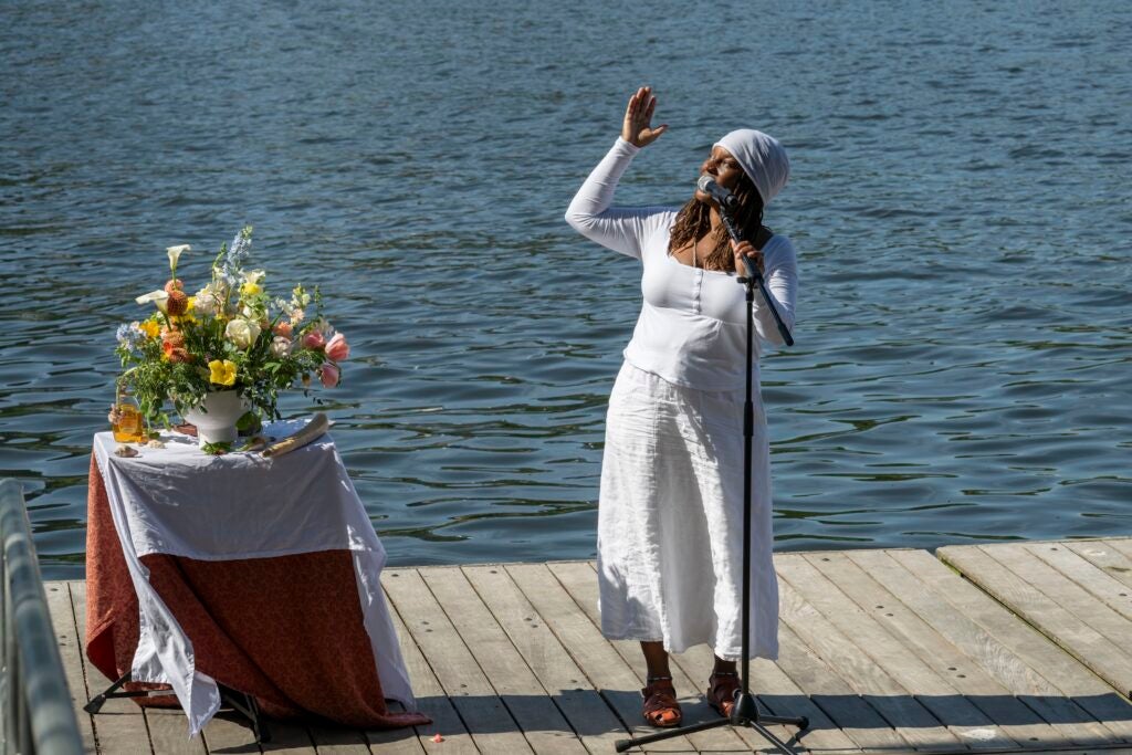 Michaela Harrison, a black woman wearing all white, stands on a wooden pier and sings into a microphone. A table with a bouquet of flowers sits beside her.